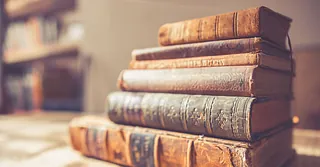 Auf einem Holztisch liegt ein Stapel alter Bücher. Die Einbände sind altersbedingt teils beschädigt, die Titel der Bücher sind nicht leserlich.