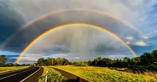 Doppelter Regenbogen über einer Landstraße mit grünen Bäumen und gelben Wiesen unter bewölktem Himmel.